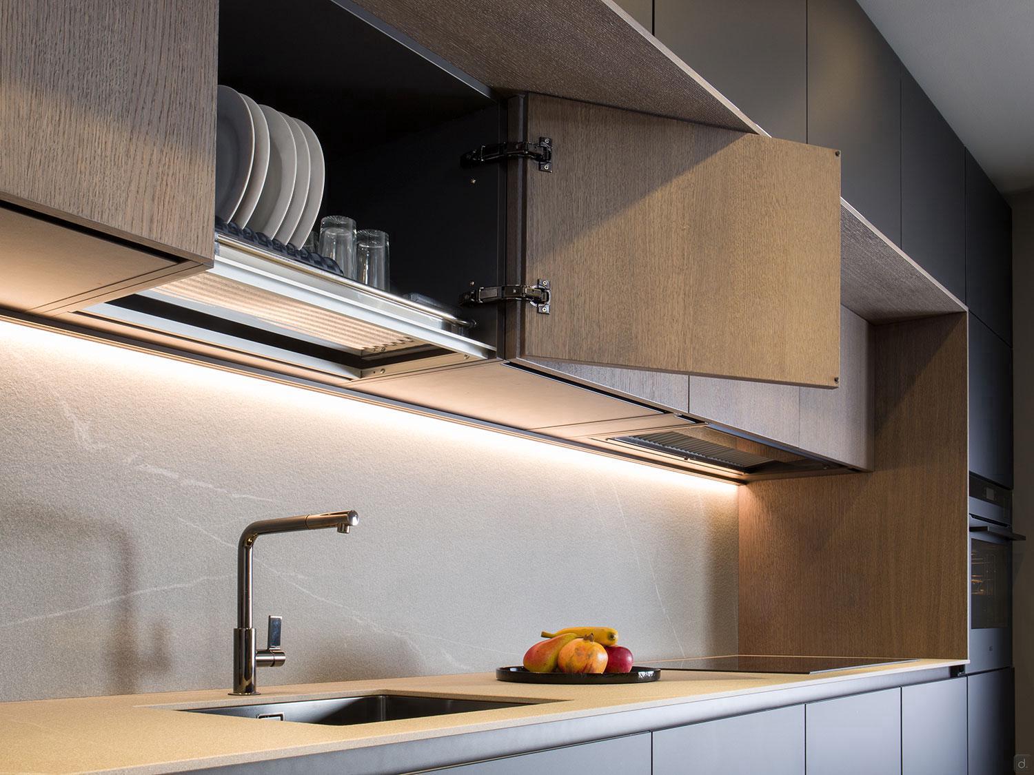 Modern Kitchens by ORDER Kitchen Furniture features light wood cabinets (some open to show plates and glasses), under-cabinet lighting illuminating a beige countertop with sink, faucet, and a bowl of fruit—perfect custom furniture for contemporary homes.