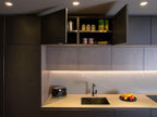 A Modern Kitchens setup by ORDER Kitchen Furniture features dark cabinets, custom storage with open cupboards displaying canned goods and containers, a sink beneath, plus a black toaster and a bowl of fruit on the beige countertop.