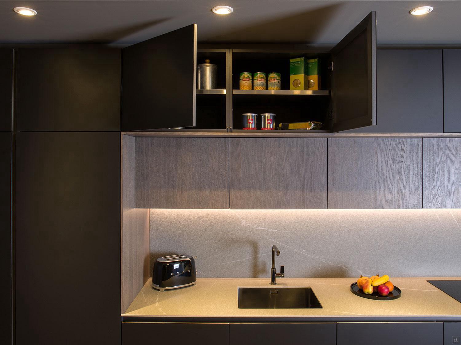 A Modern Kitchens setup by ORDER Kitchen Furniture features dark cabinets, custom storage with open cupboards displaying canned goods and containers, a sink beneath, plus a black toaster and a bowl of fruit on the beige countertop.