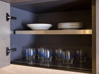 A Modern Kitchens unit by ORDER Kitchen Furniture in a Yerevan kitchen has open doors with metal hinges, showing two shelves: white plates and bowls stacked above, and eight blue-accented glasses below.