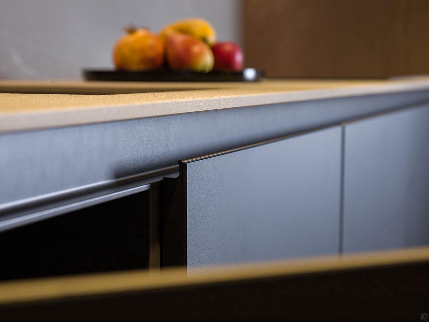 Close-up of an ORDER Kitchen Furniture matte-finish cabinet door slightly open in a Modern Kitchens setup. Above, a tray on the countertop displays assorted fruits, like apples and persimmon—ideal for stylish contemporary spaces.