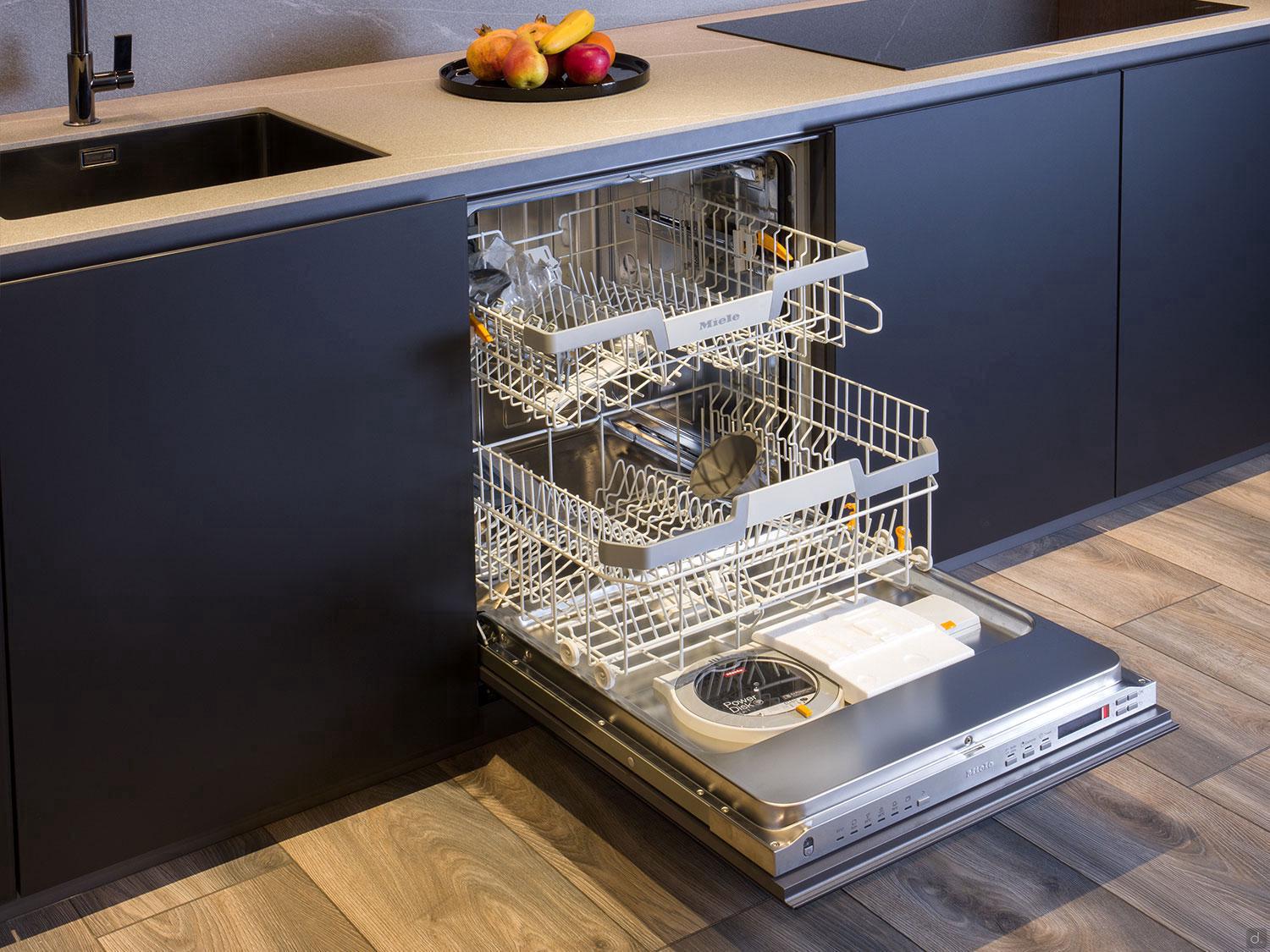 A Modern Kitchens built-in dishwasher by ORDER Kitchen Furniture stands open with empty racks, set in custom dark cabinets and wood flooring, topped with a bowl of assorted fruit near the sink.