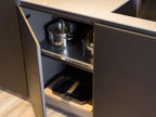 An open kitchen cabinet from ORDER Kitchen Furniture’s Modern Kitchens line displays two stainless steel pots and a grill pan on a cutting board, highlighting sleek modern functionality. The countertop and sink are also partly visible.