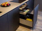 A Modern Kitchens setup by ORDER Kitchen Furniture in Yerevan features dark cabinets, custom units, open drawers with organized utensils, oven mitts, a crockpot, yellow container, and a black plate of assorted fruits on the countertop above a light wooden floor.