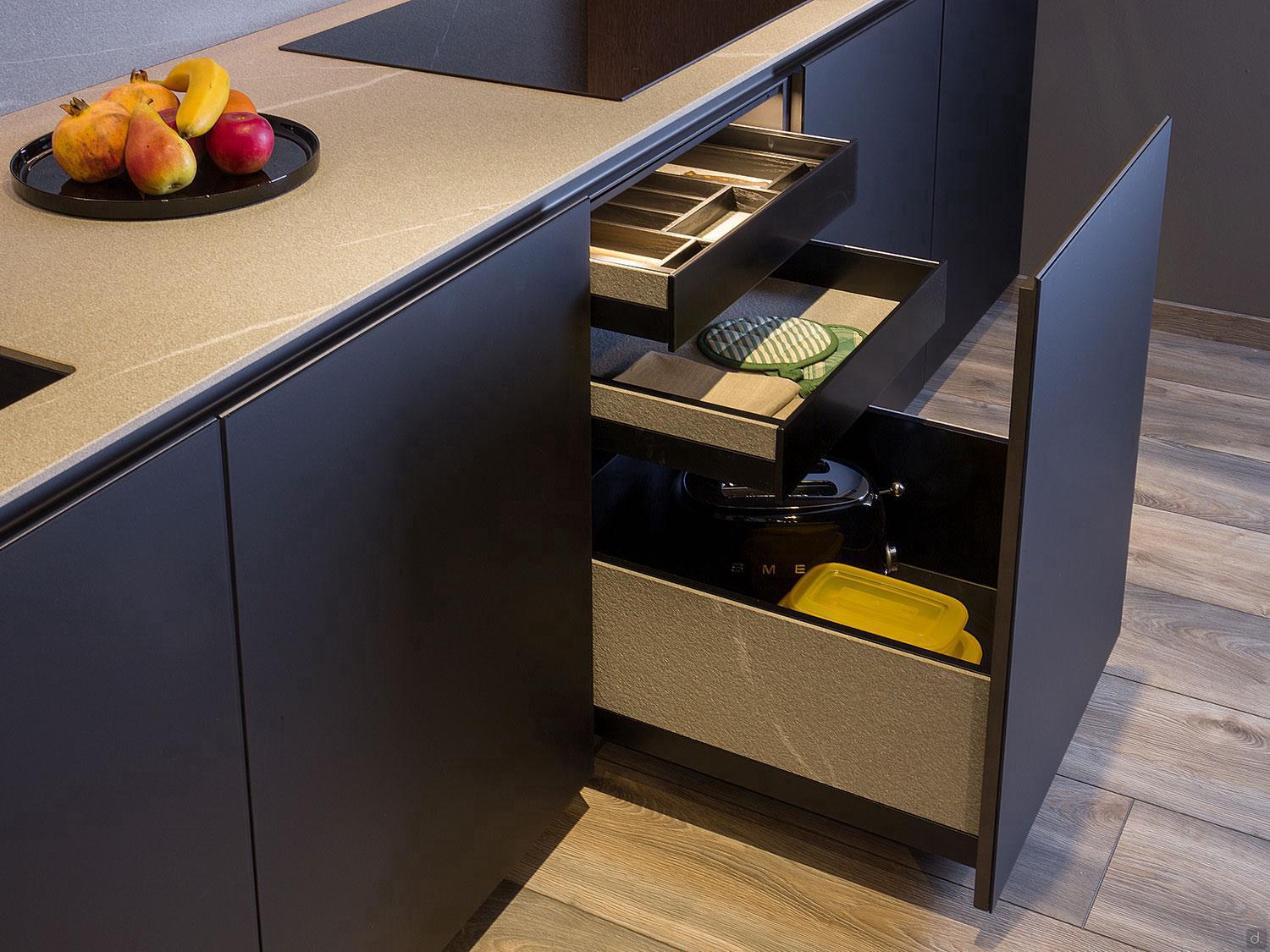 A Modern Kitchens setup by ORDER Kitchen Furniture in Yerevan features dark cabinets, custom units, open drawers with organized utensils, oven mitts, a crockpot, yellow container, and a black plate of assorted fruits on the countertop above a light wooden floor.