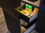 An open kitchen drawer stocked with pasta boxes, cans, and snacks highlights the sleek design of Modern Kitchens by ORDER Kitchen Furniture. Dark cabinetry and warm wood flooring complete the contemporary look.