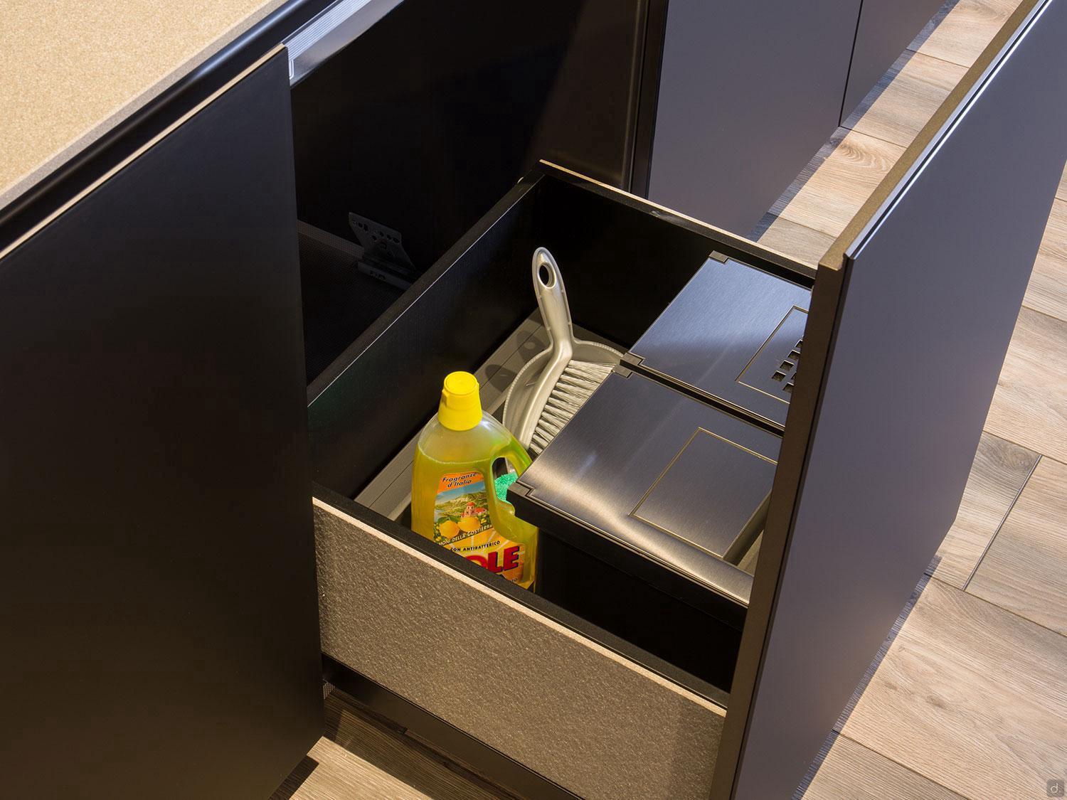 An open drawer in a Modern Kitchens design by ORDER Kitchen Furniture reveals a cleaning liquid, dustpan, brush, and two metal trash bins. The custom unit has dark finishes and rests on a sleek wooden floor.