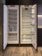Two open, empty refrigerators with glass shelves and clear drawers stand side by side in a Modern Kitchens setup by ORDER Kitchen Furniture, featuring wooden flooring and dark custom cabinetry.
