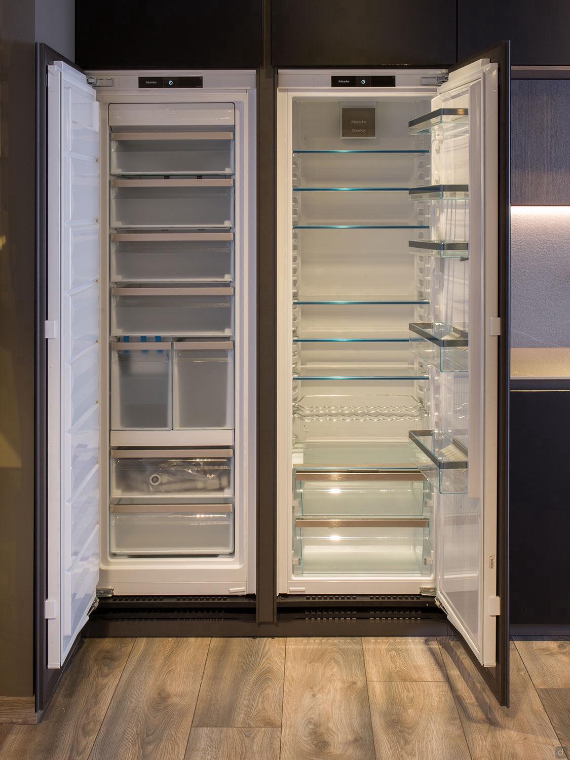 Two open, empty refrigerators with glass shelves and clear drawers stand side by side in a Modern Kitchens setup by ORDER Kitchen Furniture, featuring wooden flooring and dark custom cabinetry.