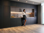 A woman in a white blouse and black skirt leans against the counter in a modern Yerevan kitchen by ORDER Kitchen Furniture, featuring dark cabinets, wood flooring, recessed lighting, and elegant Modern Kitchens design.