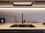 The Modern Kitchens countertop by ORDER Kitchen Furniture features custom cabinetry, a stainless steel sink and faucet, a black toaster on the left, and under-cabinet lighting highlighting the light gray textured backsplash.