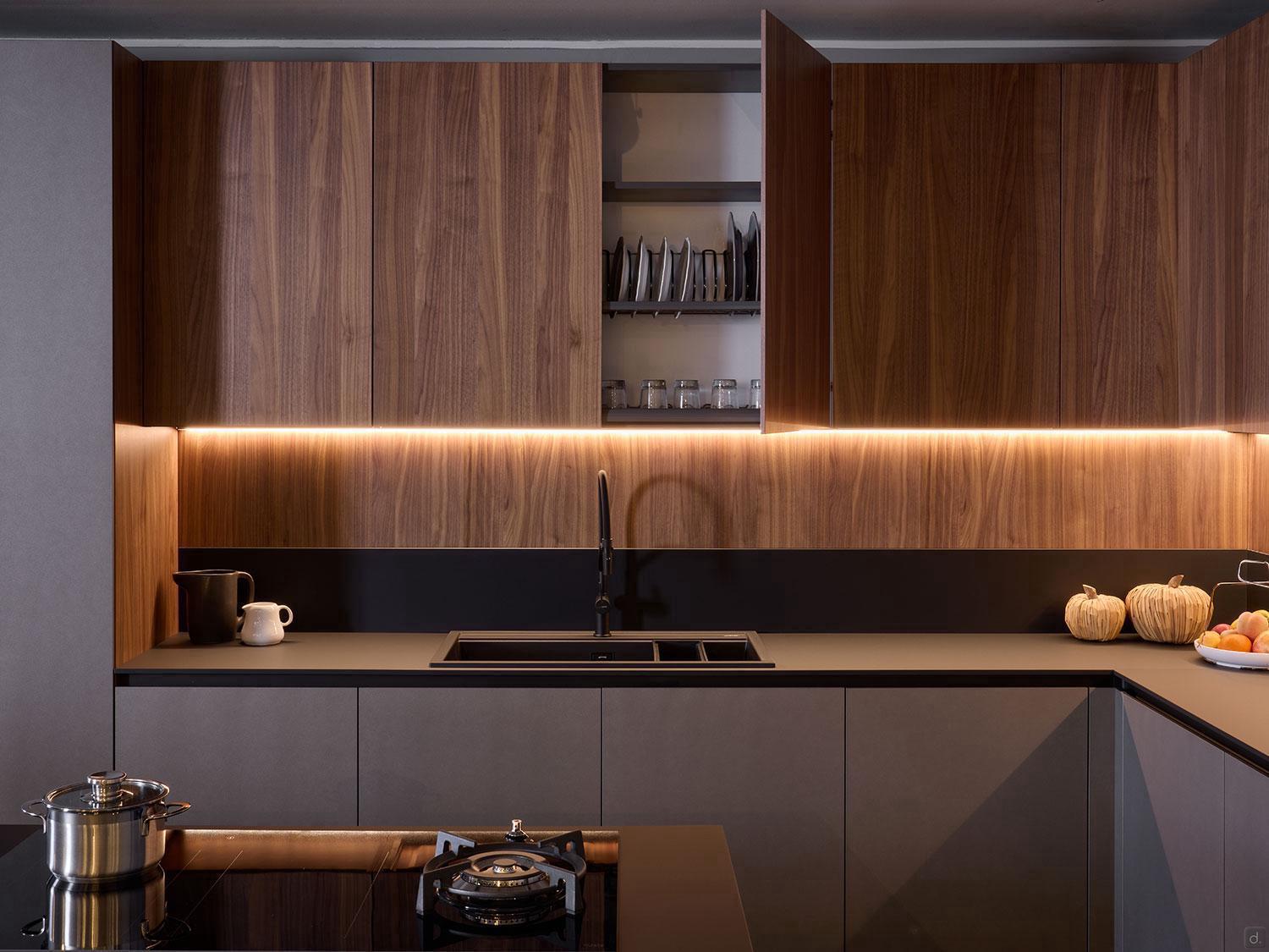 A modern kitchen by Kitchen Furniture features custom wood cabinets, some open to display dishes, with warm under-cabinet lighting. The Modern Kitchens design includes a black sink, teapot, pumpkins on the counter, and a pot on the stove in front.