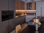 The Modern Kitchens by Kitchen Furniture features matte gray and wood cabinets, custom furniture, built-in black ovens, under-cabinet lighting, a dark island with a vase of flowers and pumpkins, a beige chair, and a stovetop pot.