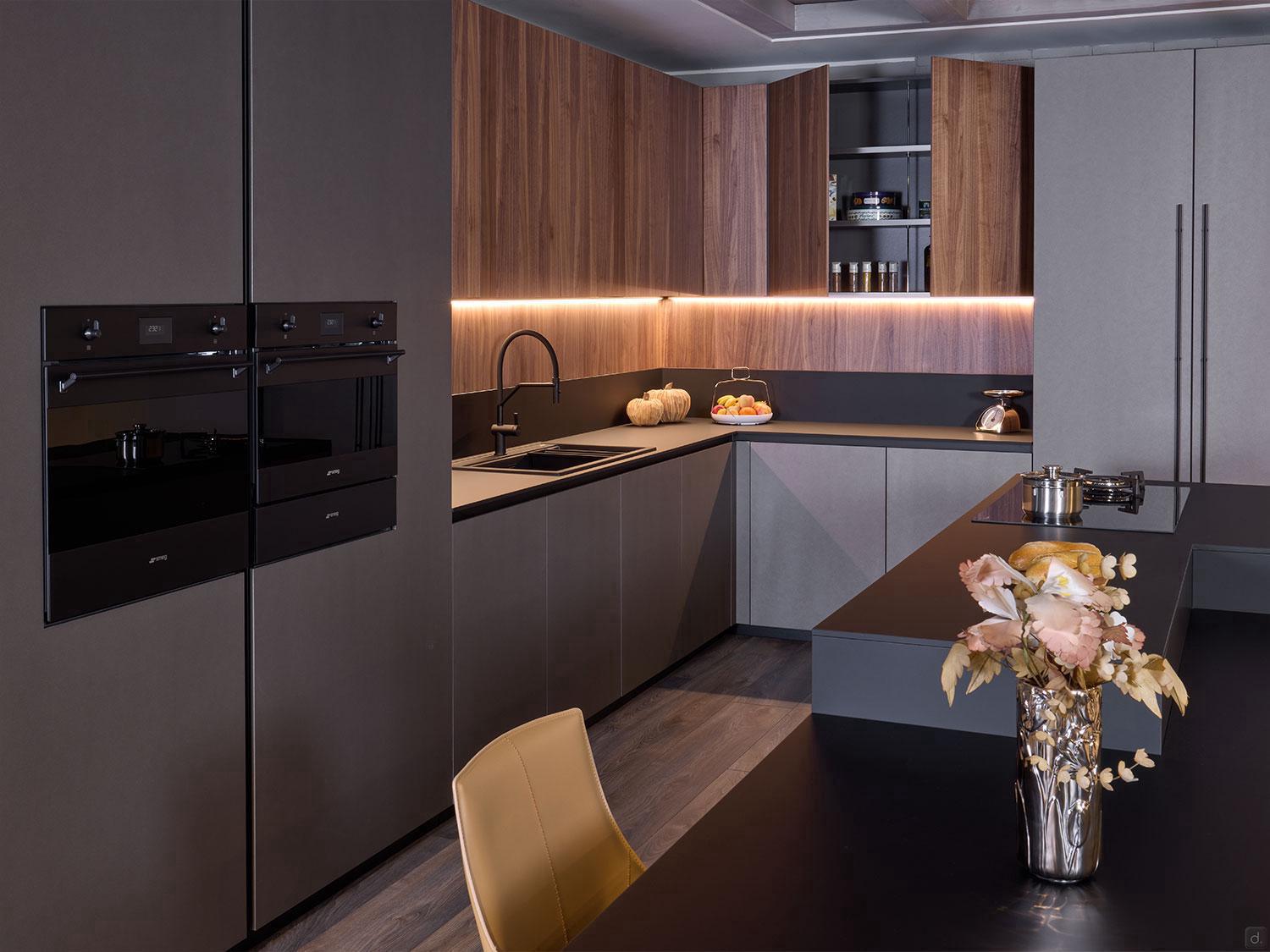 The Modern Kitchens by Kitchen Furniture features matte gray and wood cabinets, custom furniture, built-in black ovens, under-cabinet lighting, a dark island with a vase of flowers and pumpkins, a beige chair, and a stovetop pot.