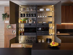 Modern Kitchens by Kitchen Furniture features open cabinets with dishes and glasses, two built-in ovens, a wine rack, small appliances, and a black counter displaying a fruit bowl with bananas, apples, and grapes.