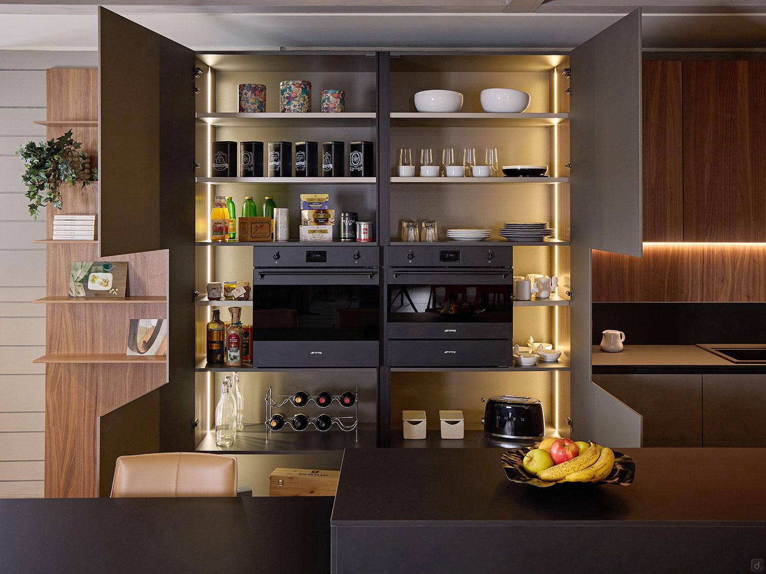 Modern Kitchens by Kitchen Furniture features open cabinets with dishes and glasses, two built-in ovens, a wine rack, small appliances, and a black counter displaying a fruit bowl with bananas, apples, and grapes.