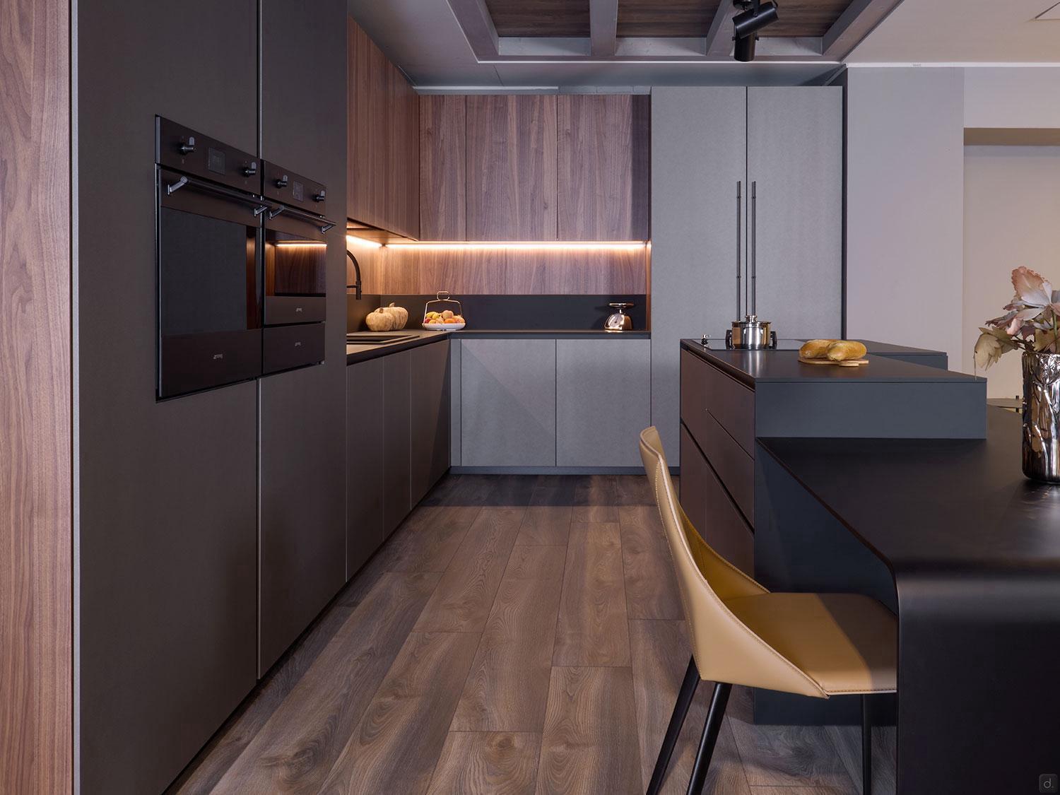 The Modern Kitchens by Kitchen Furniture features wood and grey cabinets, black built-in ovens, under-cabinet lighting, a dark island with beige chairs, custom cabinetry, wood flooring, and a countertop decorated with candles, fruit, and flowers.