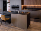 Modern Kitchens by Kitchen Furniture offers sleek cabinets, a black island cooker, built-in ovens, wood-paneled walls, under-cabinet lighting, a black sink, and a tan chair on light wood floors with custom kitchen furniture.
