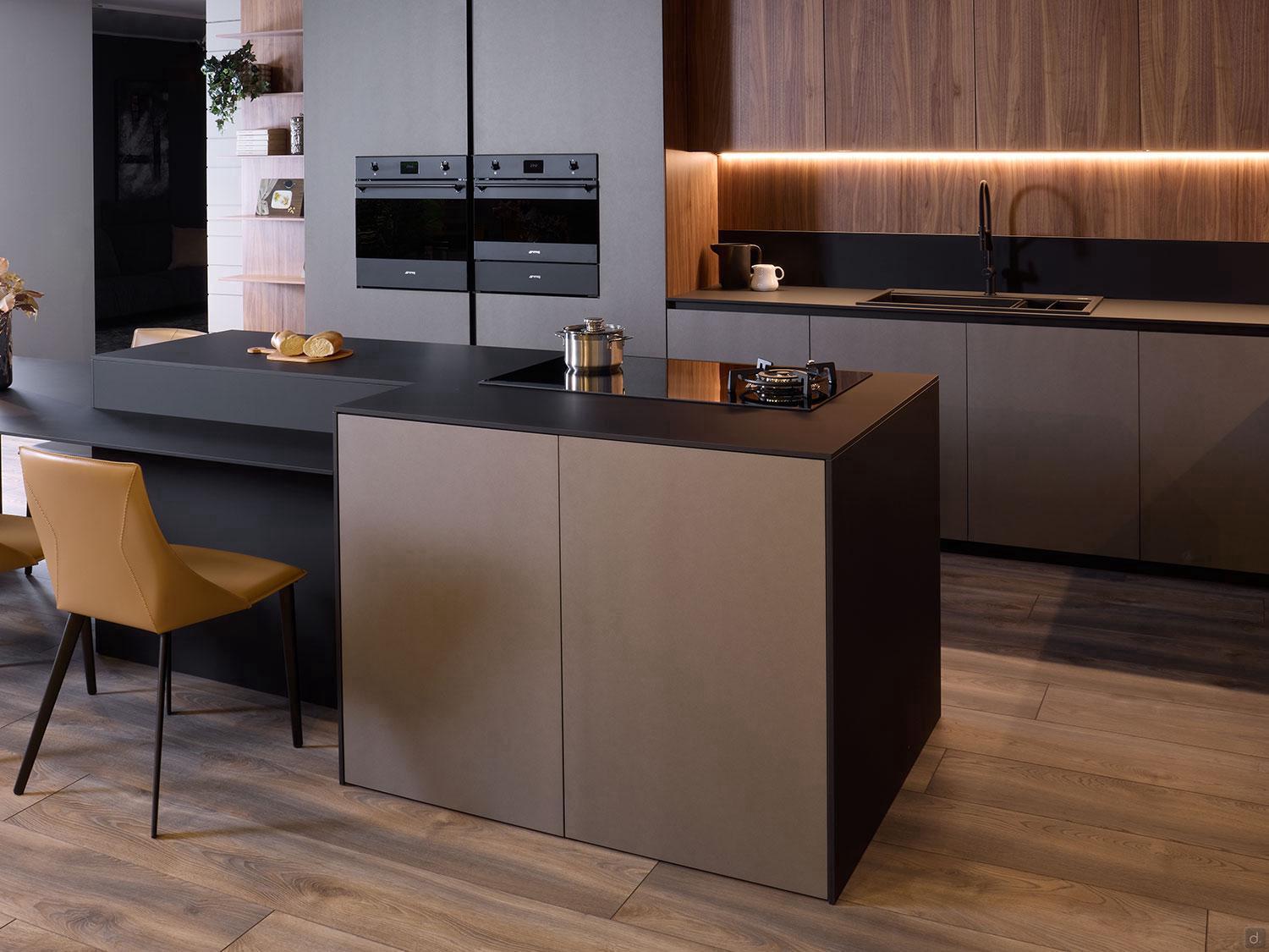 Modern Kitchens by Kitchen Furniture offers sleek cabinets, a black island cooker, built-in ovens, wood-paneled walls, under-cabinet lighting, a black sink, and a tan chair on light wood floors with custom kitchen furniture.