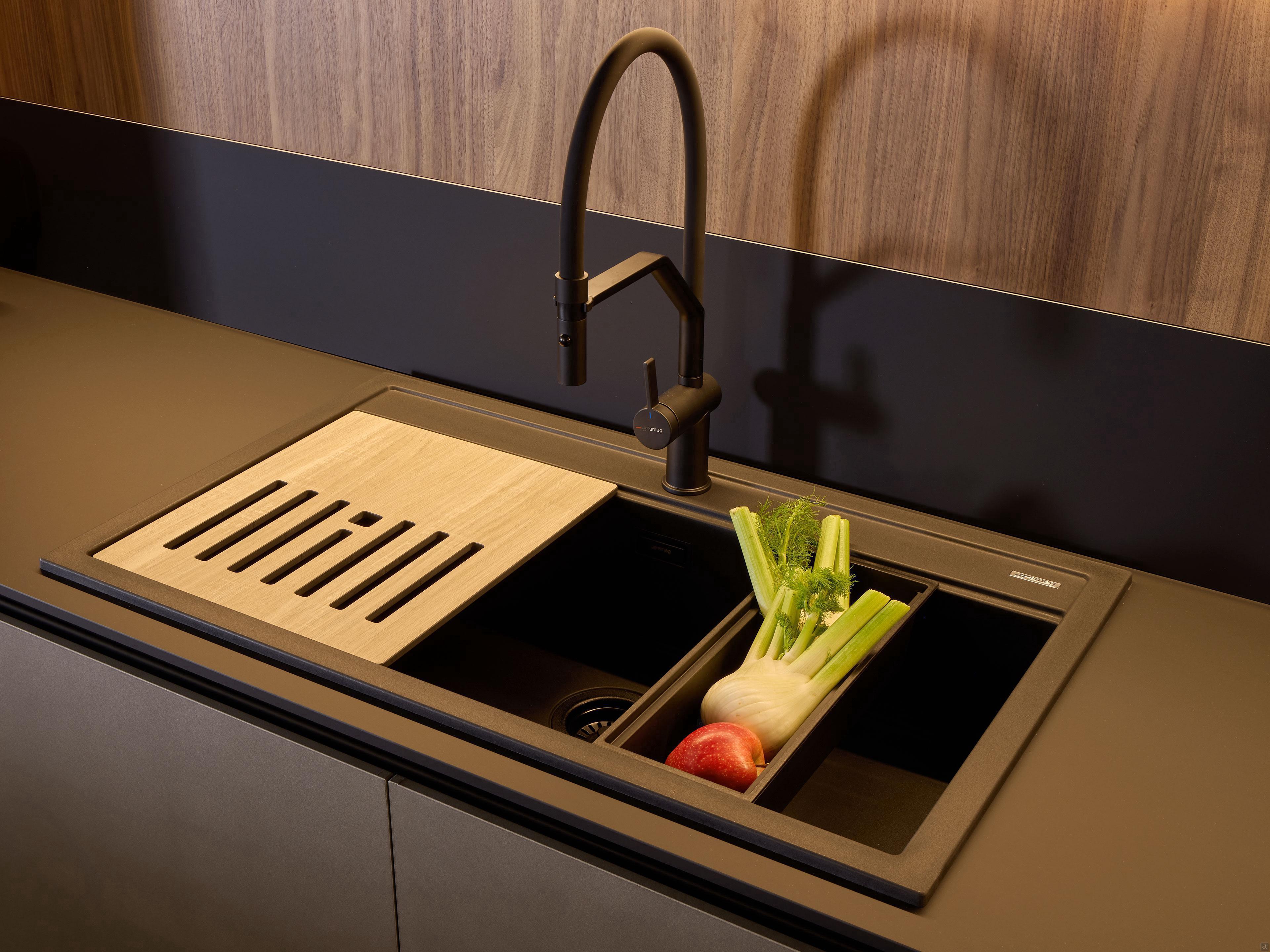 Showcasing Kitchen Furniture’s Modern Kitchens: a matte black faucet, wooden cutting board, and black container with celery, fennel, and a red apple stand out against a wood-paneled backsplash—ideal for custom kitchen design.