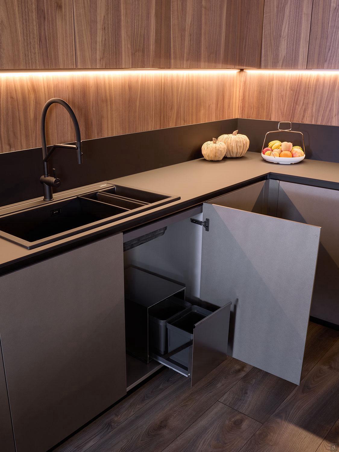The Modern Kitchens set from Kitchen Furniture features gray custom cabinets, an open corner cabinet with pull-out trash bins, a black sink and faucet, wood backsplash with under-cabinet lighting, plus a countertop displaying pumpkins and fruit.