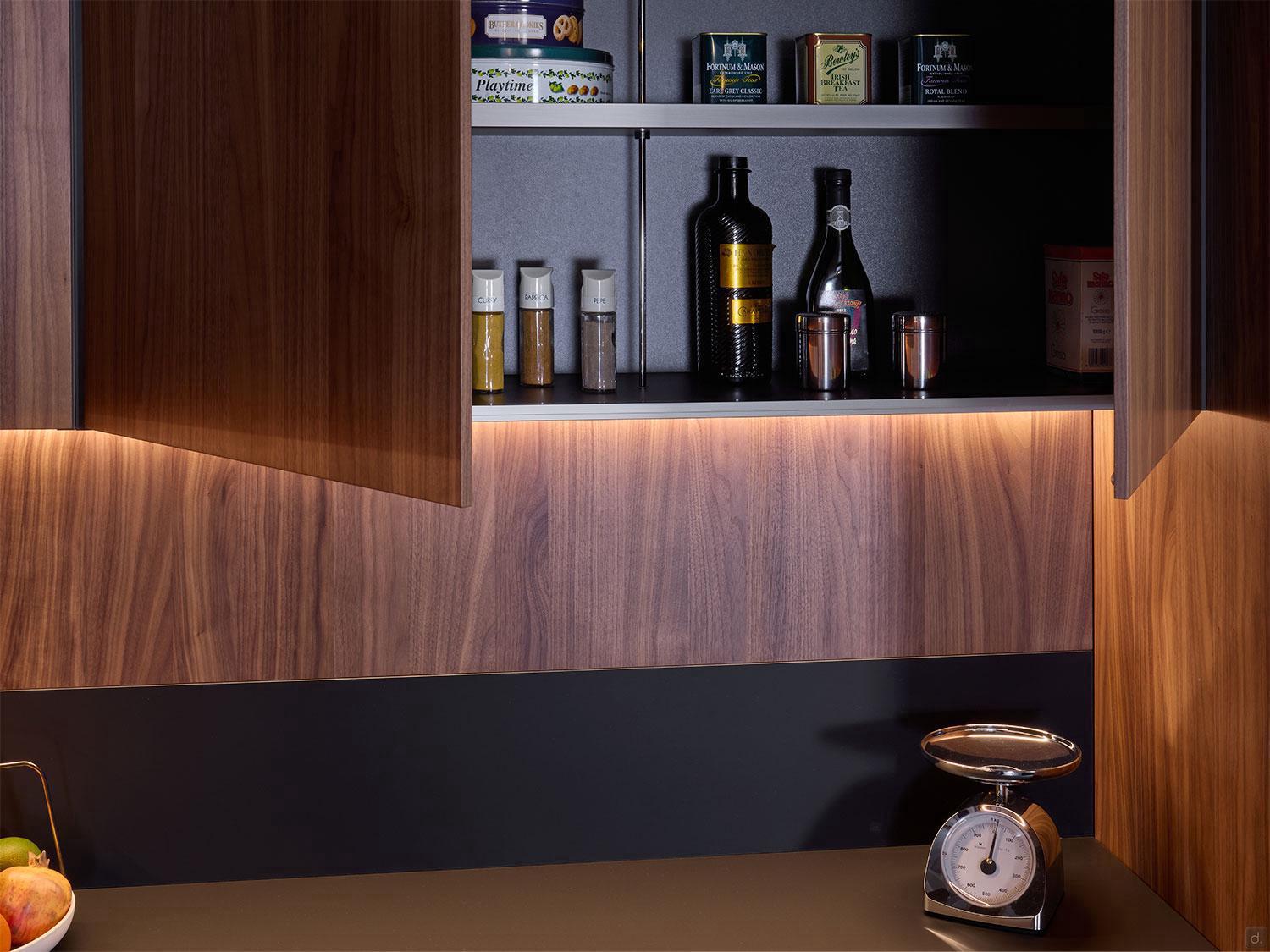 Modern Kitchens by Kitchen Furniture features a wooden cabinet with open doors displaying bottles, jars, and tins on shelves. The countertop holds fruit and a metal scale, highlighted by warm under-cabinet lighting—perfect for Yerevan homes.