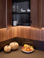 Modern Kitchens by Kitchen Furniture features dark wood cabinets revealing glassware, bowls, and plates, with a countertop showing assorted fruit and two pumpkin-shaped decorations, all highlighted by warm under-cabinet lighting.