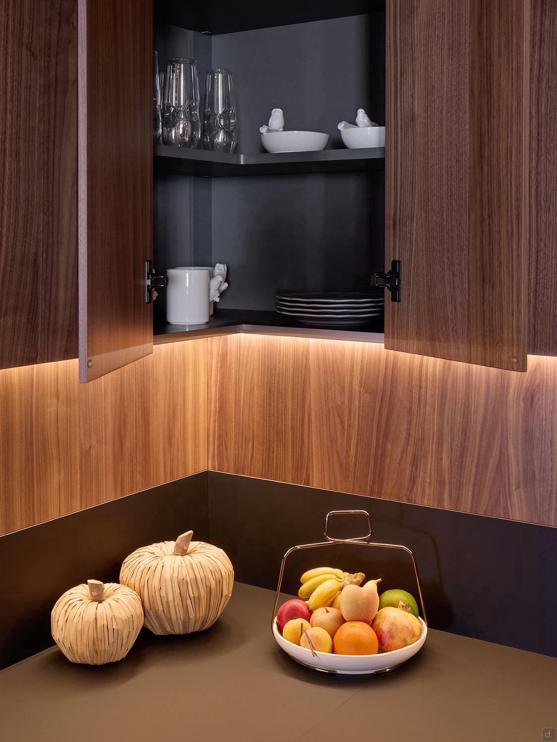 Modern Kitchens by Kitchen Furniture features dark wood cabinets revealing glassware, bowls, and plates, with a countertop showing assorted fruit and two pumpkin-shaped decorations, all highlighted by warm under-cabinet lighting.