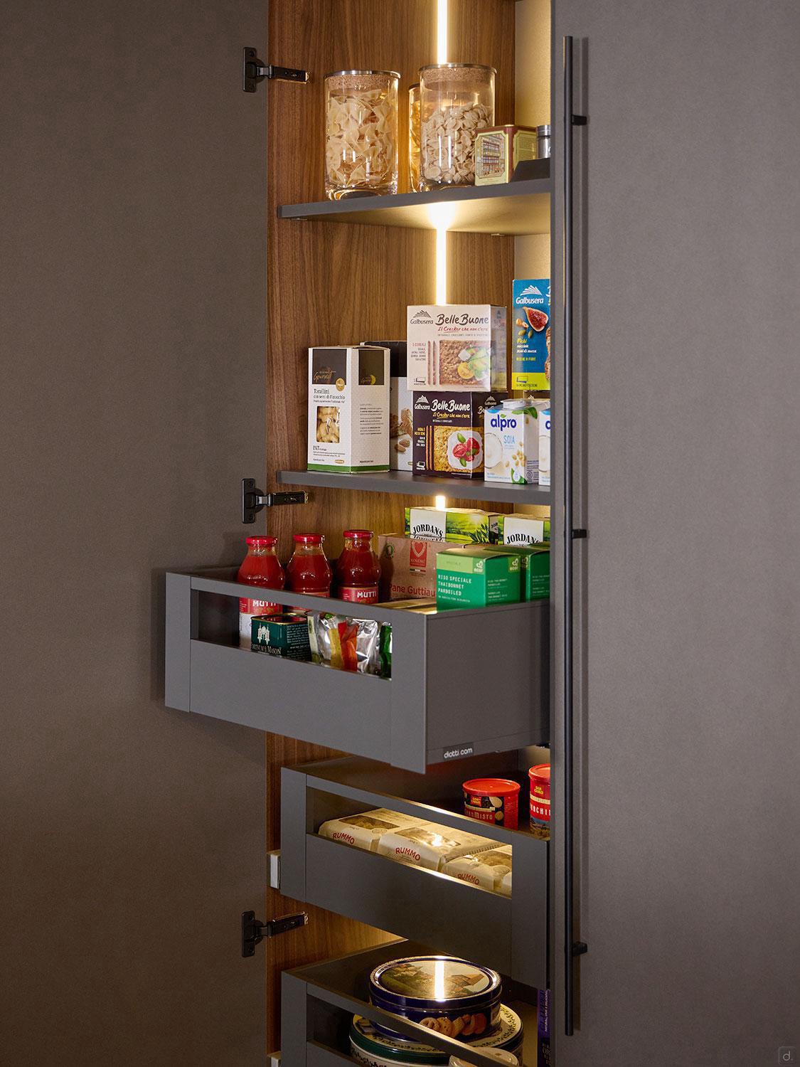 The Kitchen Furniture Modern Kitchens pantry features tall, open wooden shelves with built-in lighting, organized for jars, bottles, and groceries. Select shelves include pull-out drawers for easy access—ideal for contemporary kitchens.