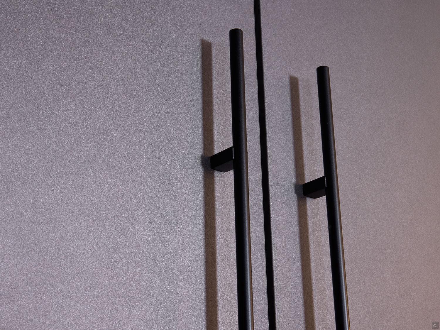 Two vertical black metal handles are mounted on a textured gray surface by Kitchen Furniture's Modern Kitchens, each casting a shadow leftward for added style—a sleek example of contemporary custom kitchen design.