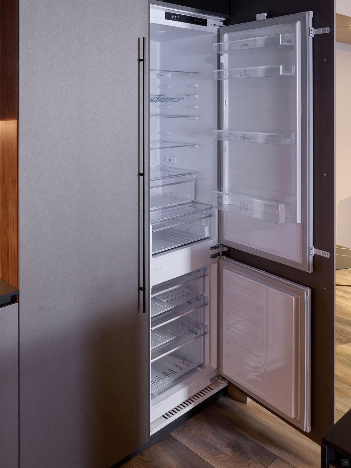 The built-in refrigerator and freezer from Kitchen Furniture’s Modern Kitchens line stand open, displaying clear shelves and drawers amid stylish wood and gray paneling, highlighting the elegance of custom kitchen furniture.