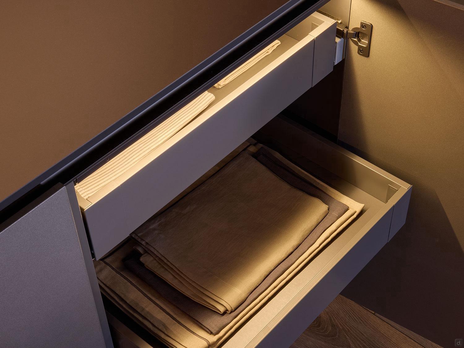 A close-up of an open Modern Kitchens drawer by Kitchen Furniture shows neatly folded beige and brown linens, highlighted by soft lighting—an elegant fit for any brown-toned interior.