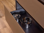 A drawer from the Modern Kitchens collection by Kitchen Furniture opens in a stylish kitchen, revealing two stainless steel pots inside. The custom brown cabinetry and light wood flooring enhance the contemporary design.