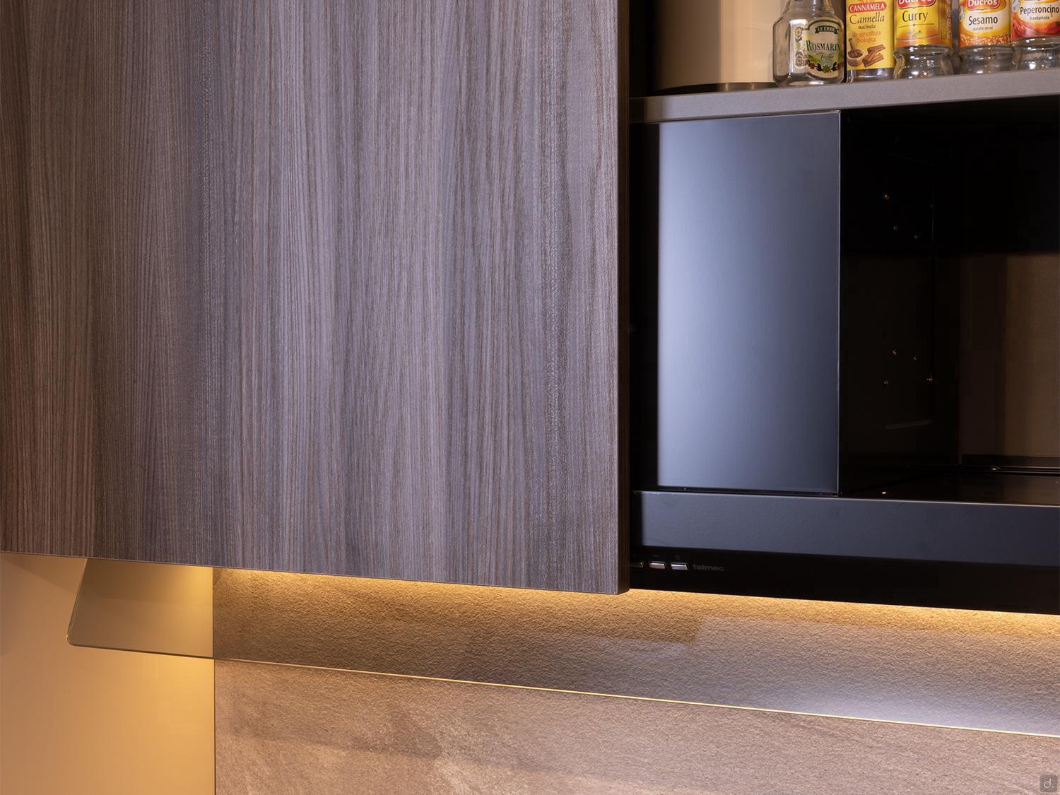 A close-up of a Modern Kitchens setup by Kitchen Furniture in Yerevan features wood-textured cabinets, an open black microwave, spice jars on top, and warm under-cabinet lighting highlighting the textured backsplash.