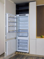 A modern built-in refrigerator from Kitchen Furniture’s Modern Kitchens collection stands open, displaying empty shelves, drawers, and a wine rack beside sleek custom kitchen furniture and countertops.