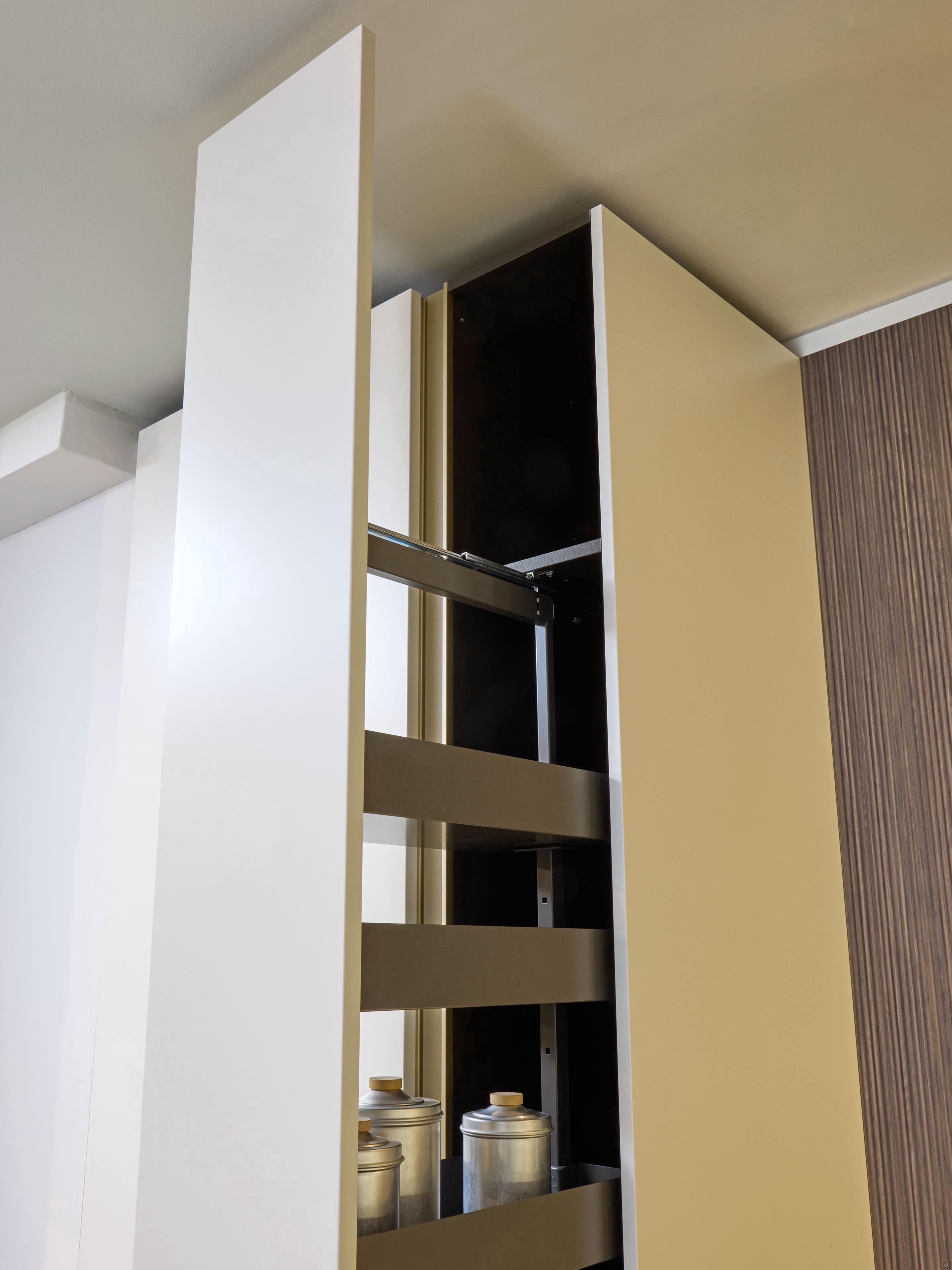The Modern Kitchens by Kitchen Furniture features a tall, sleek cabinet with a pull-out vertical pantry, partially open to show shelves with metal canisters and wooden lids—perfect for enhancing any neutral-toned, minimalist kitchen design.