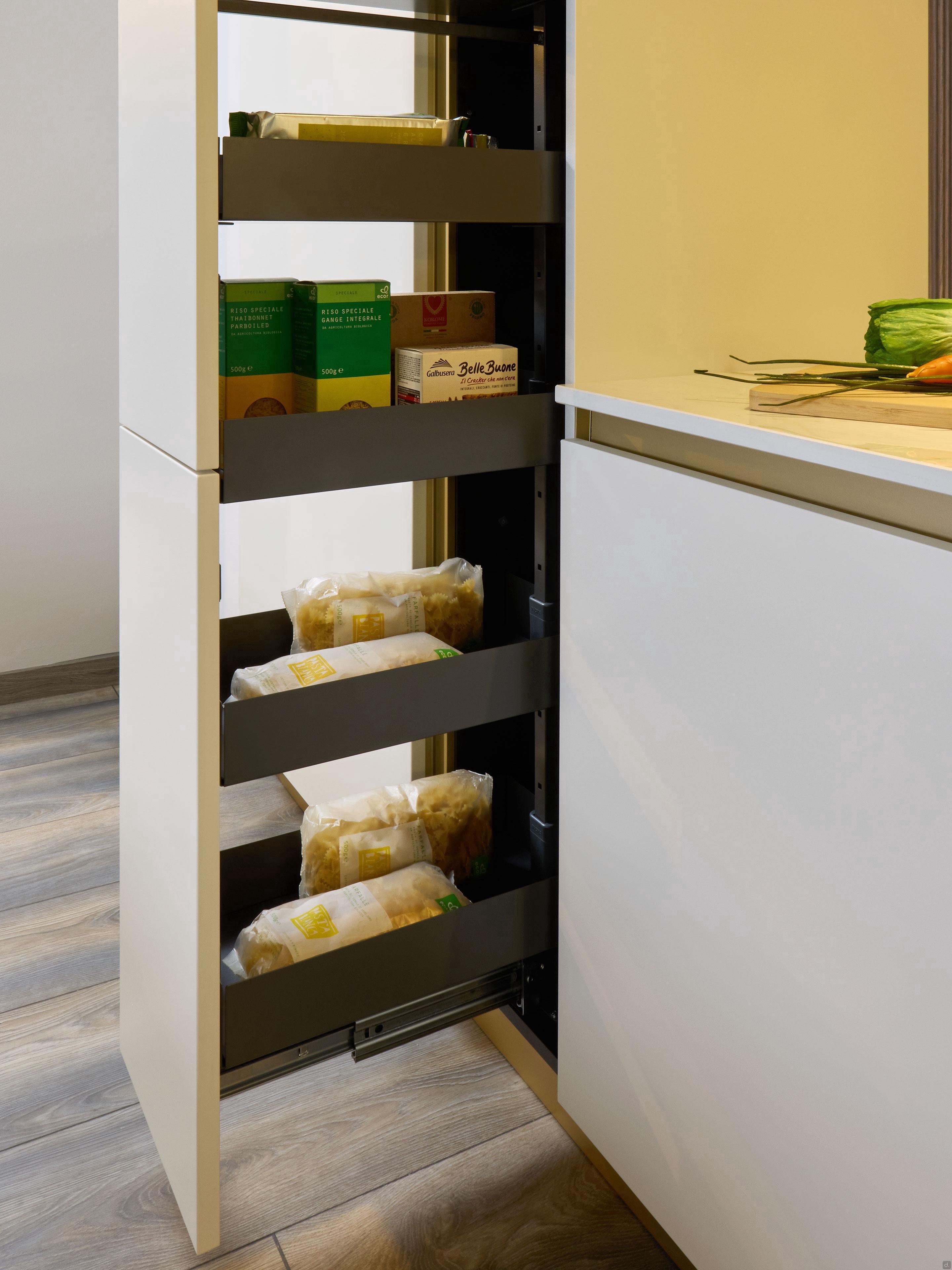 A Modern Kitchens pull-out pantry cabinet by Kitchen Furniture, partially open beside a white countertop with sliced zucchini, displays boxes of dry food and pasta—highlighting sleek, custom kitchen design.