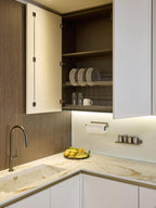 Modern Kitchens by Kitchen Furniture showcases an open upper cabinet with plates, bowls, and glasses on wire racks, a marble countertop with fruit bowl, sink, paper towel holder, spice jars, and sleek under-cabinet lighting.