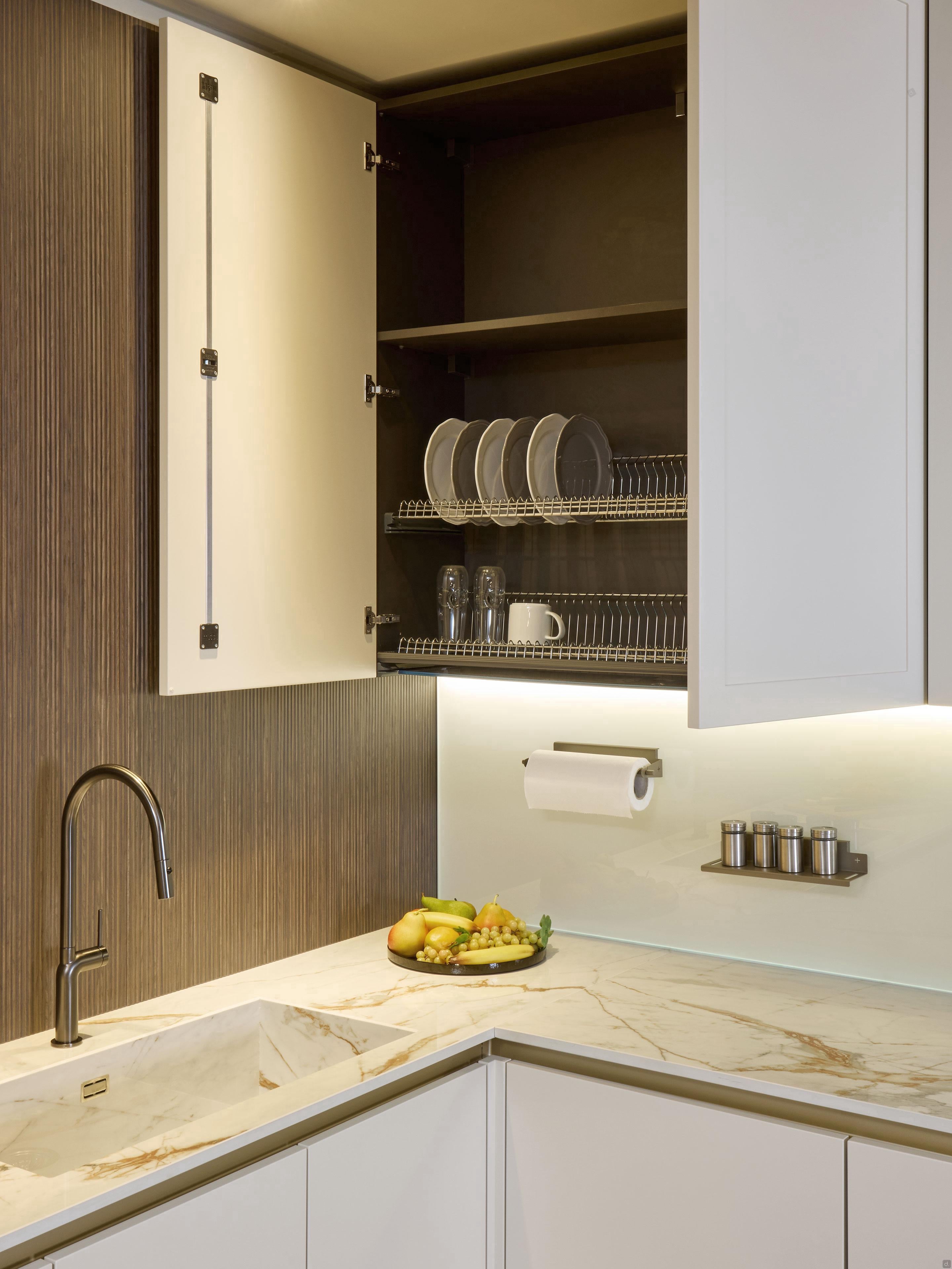 Modern Kitchens by Kitchen Furniture showcases an open upper cabinet with plates, bowls, and glasses on wire racks, a marble countertop with fruit bowl, sink, paper towel holder, spice jars, and sleek under-cabinet lighting.