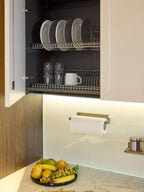A kitchen cabinet with open doors displays plates, glasses, and a mug on drying racks. Below, a wall-mounted paper towel roll and a bowl of fruit sit on marble—showcasing Kitchen Furniture’s Modern Kitchens style.