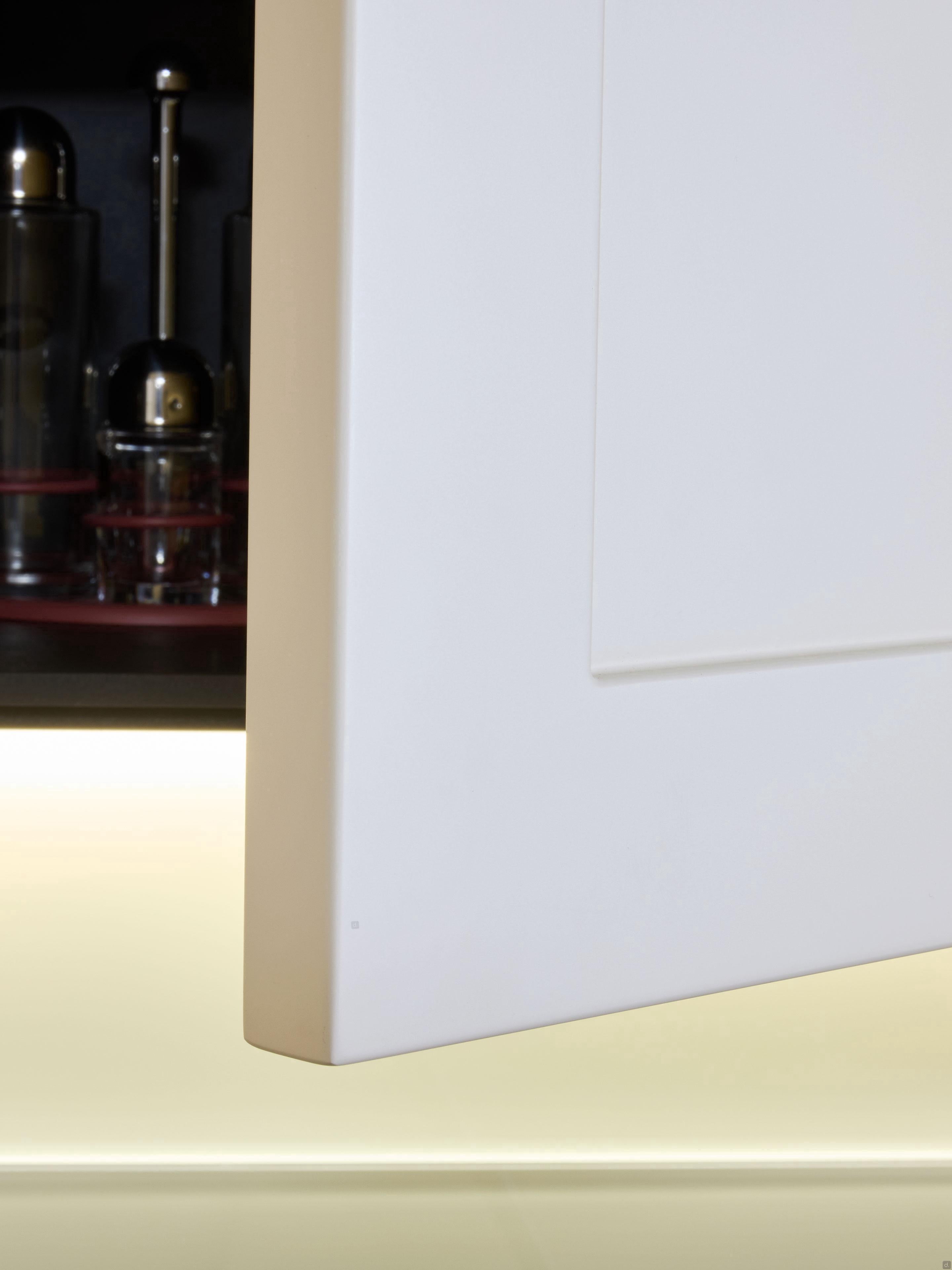 A close-up of a white cabinet door from Modern Kitchens by Kitchen Furniture, with blurred glass kitchenware and utensils on a shelf in the background.