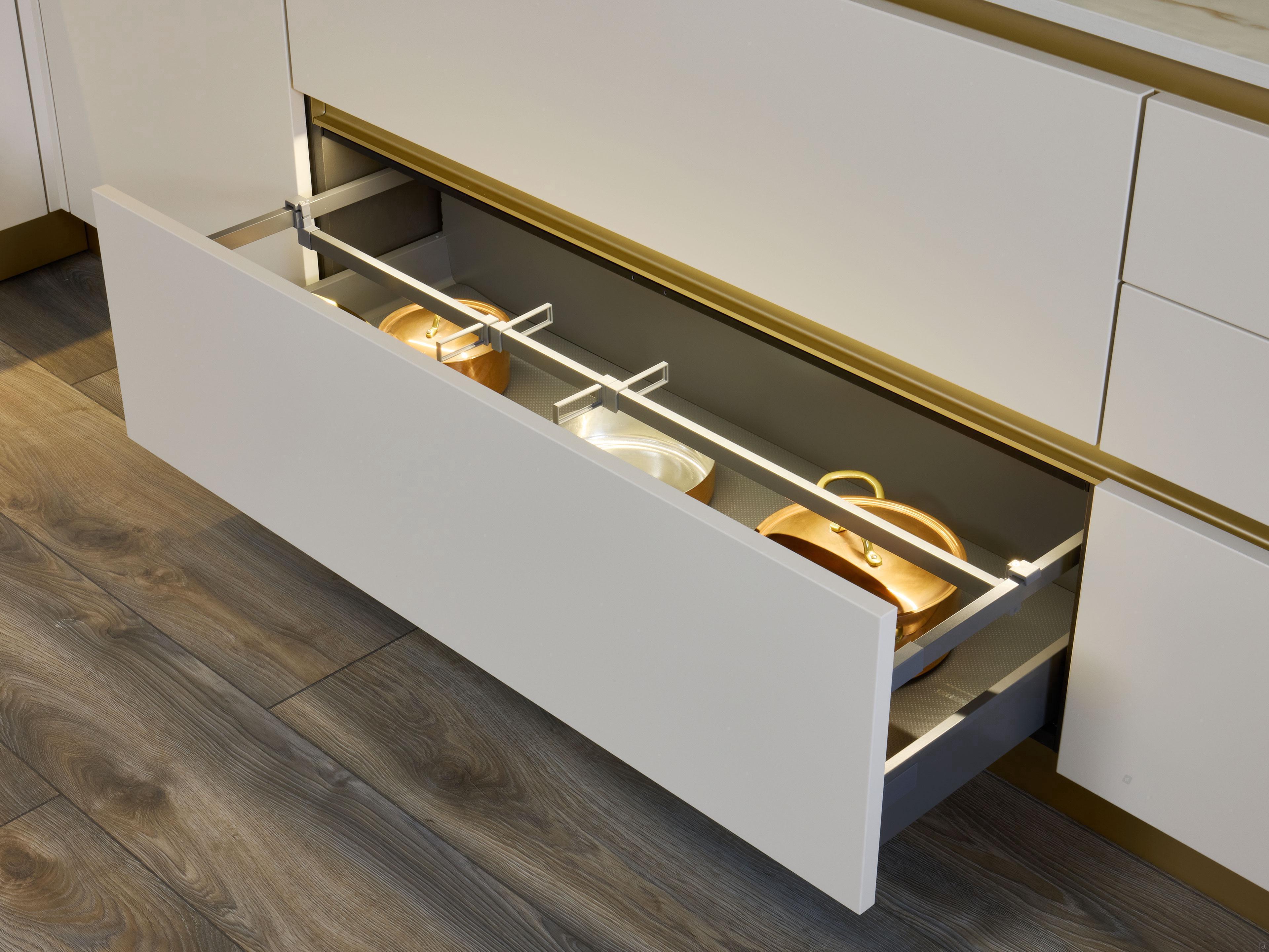 A Modern Kitchens drawer from Kitchen Furniture opens to reveal neatly organized pots and lids, showcasing custom storage solutions with gold accents and wood-grain tile flooring that enhance your kitchen’s style.