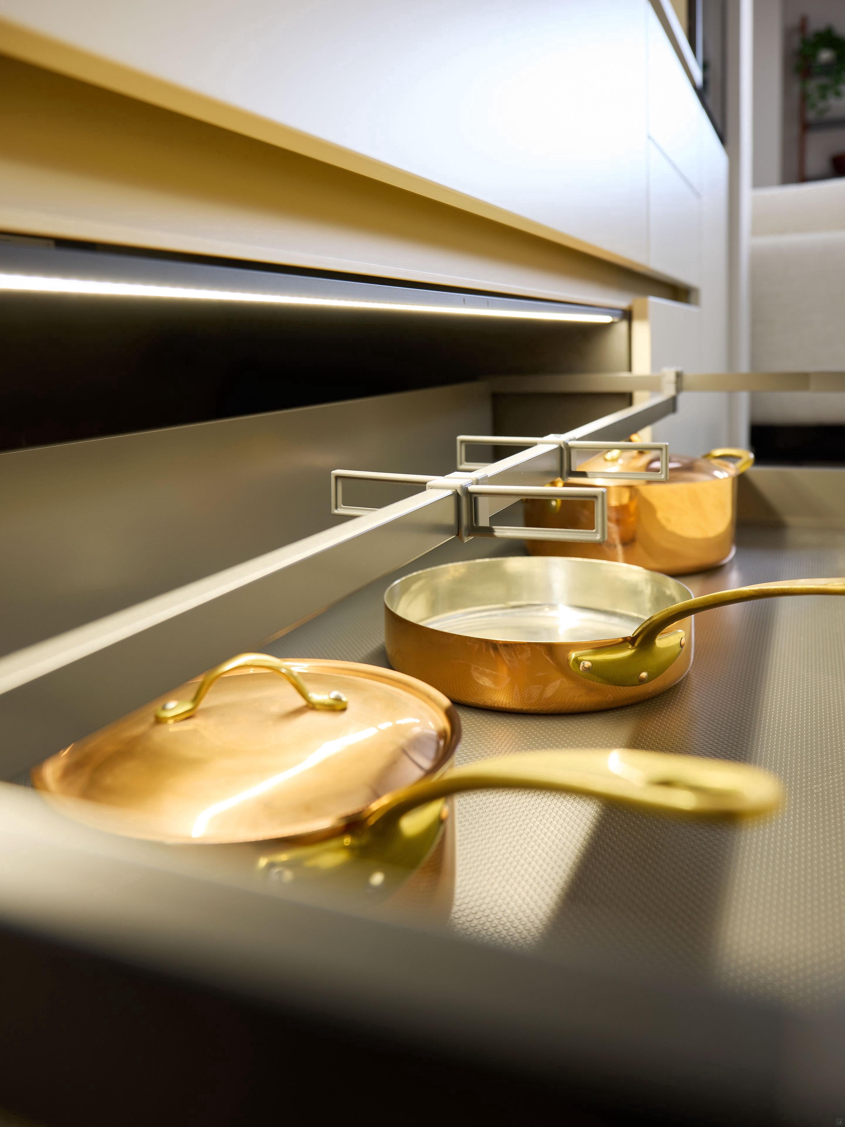 Close-up of three shiny copper pots with gold handles organized in a Modern Kitchens drawer by Kitchen Furniture, featuring a soft, neutral interior and sleek custom kitchen design.