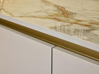 Close-up of a Modern Kitchens countertop by Kitchen Furniture, featuring polished marble with gold and brown veining above sleek white cabinets with gold accent trim and custom kitchen furniture.