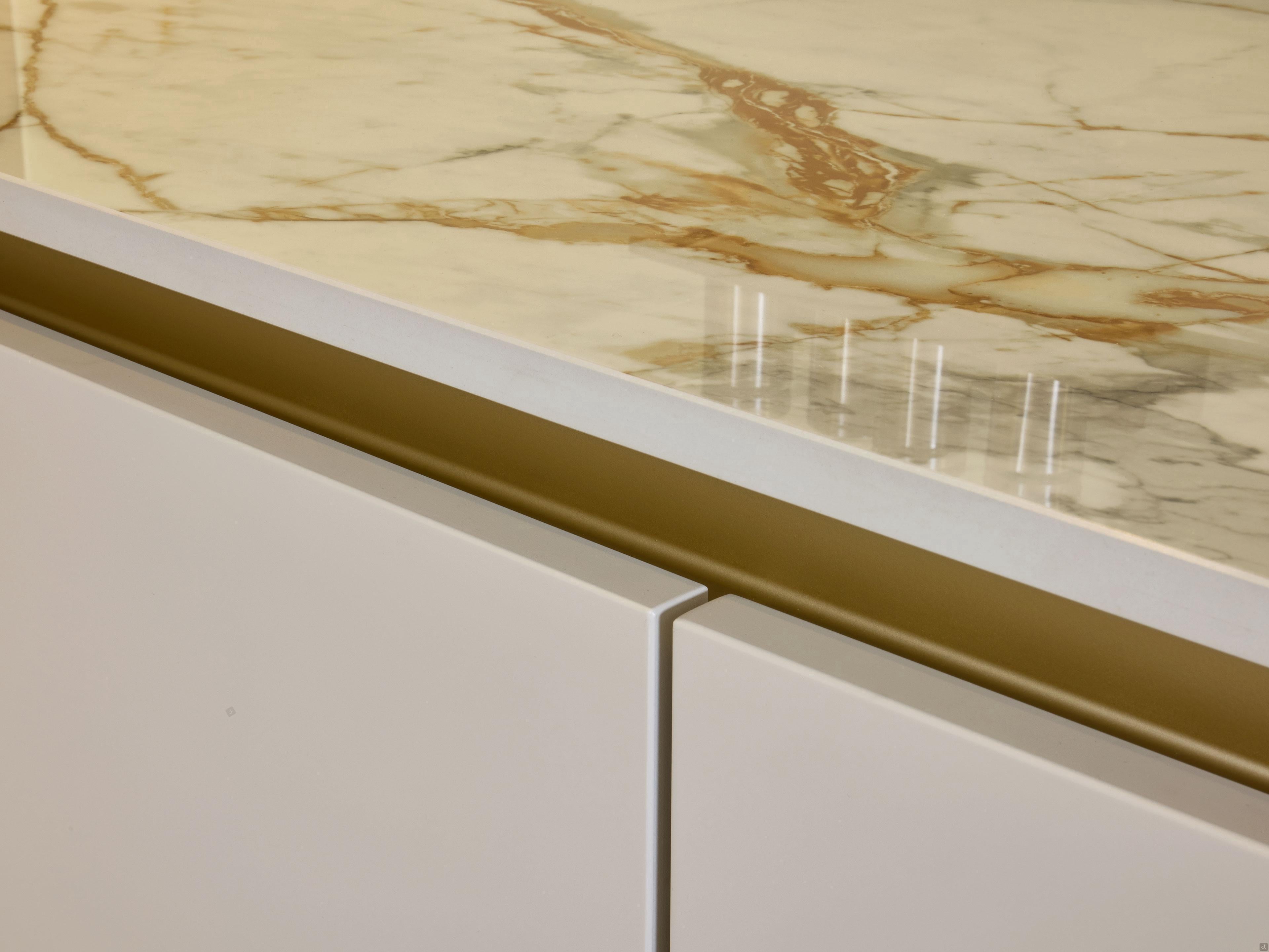 Close-up of a Modern Kitchens countertop by Kitchen Furniture, featuring polished marble with gold and brown veining above sleek white cabinets with gold accent trim and custom kitchen furniture.