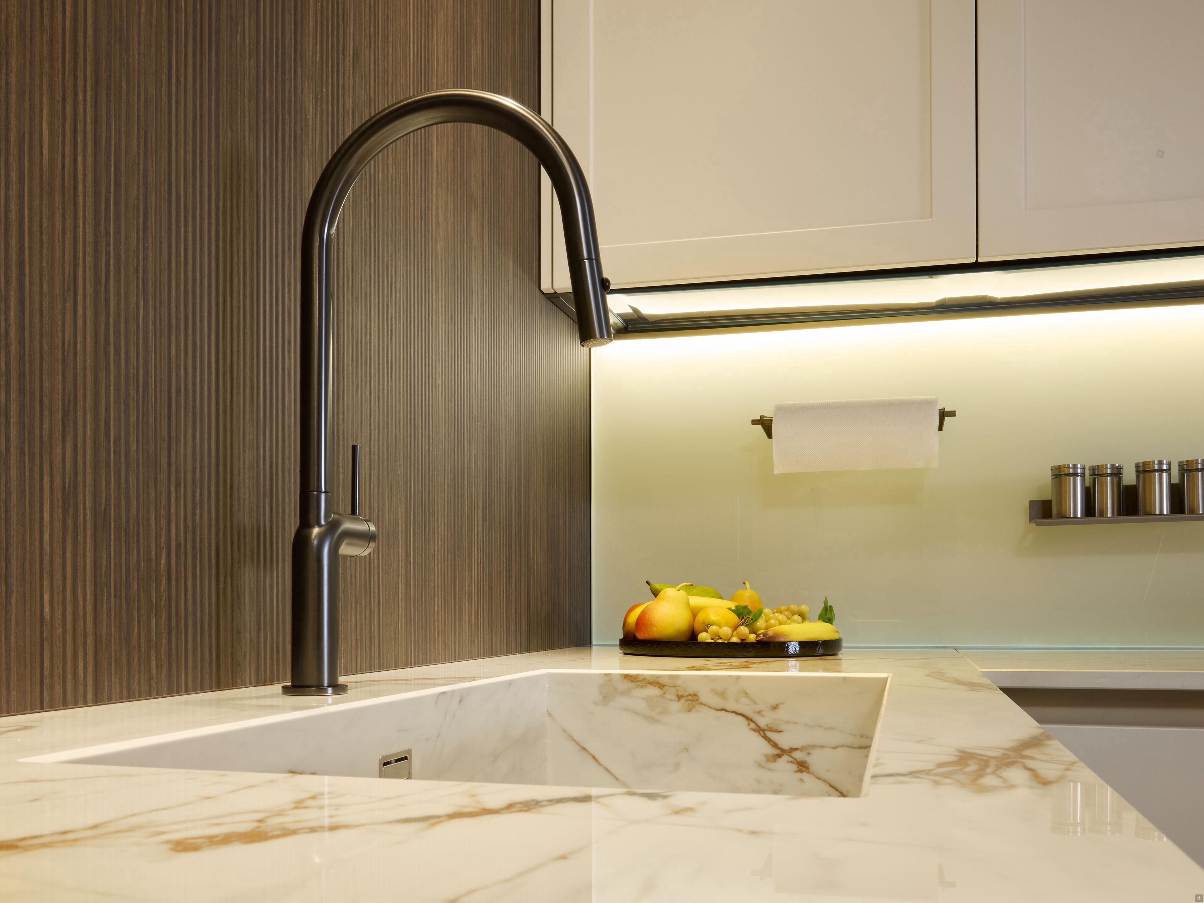The Modern Kitchens by Kitchen Furniture features a sleek black faucet on a marble countertop, white cabinets, wooden wall panel, spice jars, and fruits—creating a stylish, refined modern kitchen Yerevan aesthetic.
