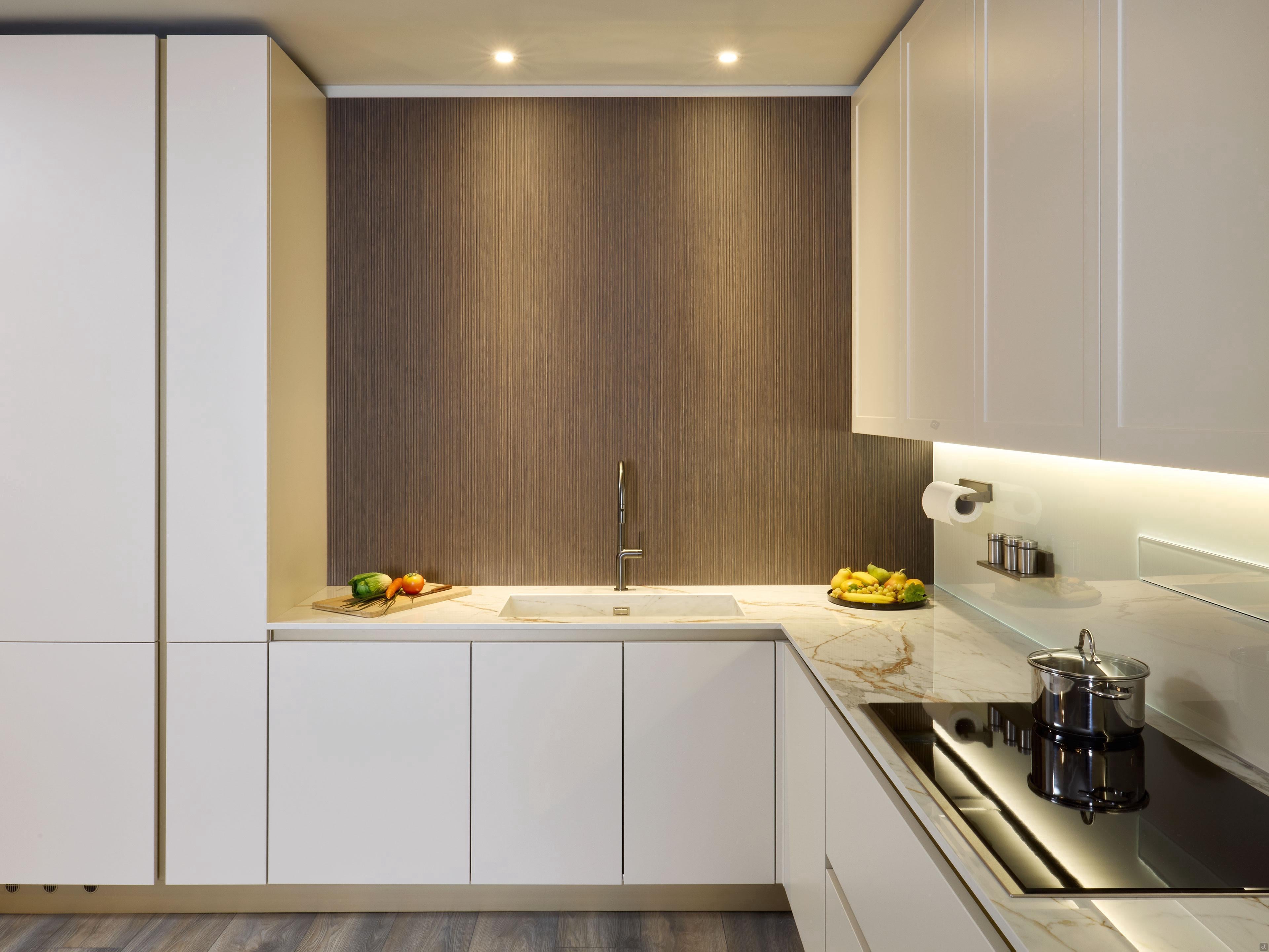 Modern Kitchens by Kitchen Furniture in Yerevan features white cabinets, marble countertop, electric stove, tall faucet, under-cabinet lighting, wooden accent wall, plus fresh veggies and a fruit bowl for a stylish touch.