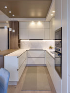 Modern Kitchens by Kitchen Furniture features white cabinets, a built-in black oven, marble backsplash, under-cabinet lighting, wooden floors with a beige rug, pendant lights over the countertop, and custom kitchen furniture.