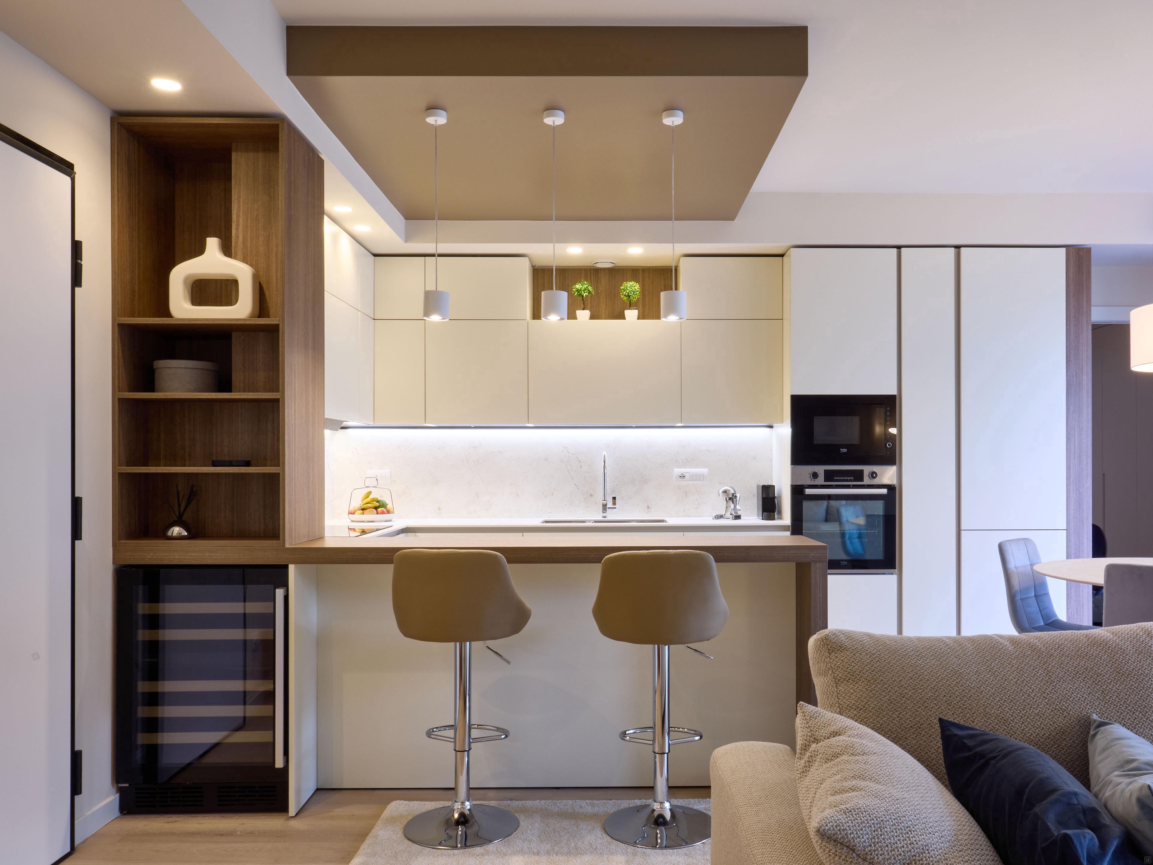 Modern Kitchens by Kitchen Furniture features white cabinets, wood accents, built-in oven, under-cabinet lighting, two beige bar stools, pendant lights above the counter, open shelving with decor, and a wine fridge on the left.