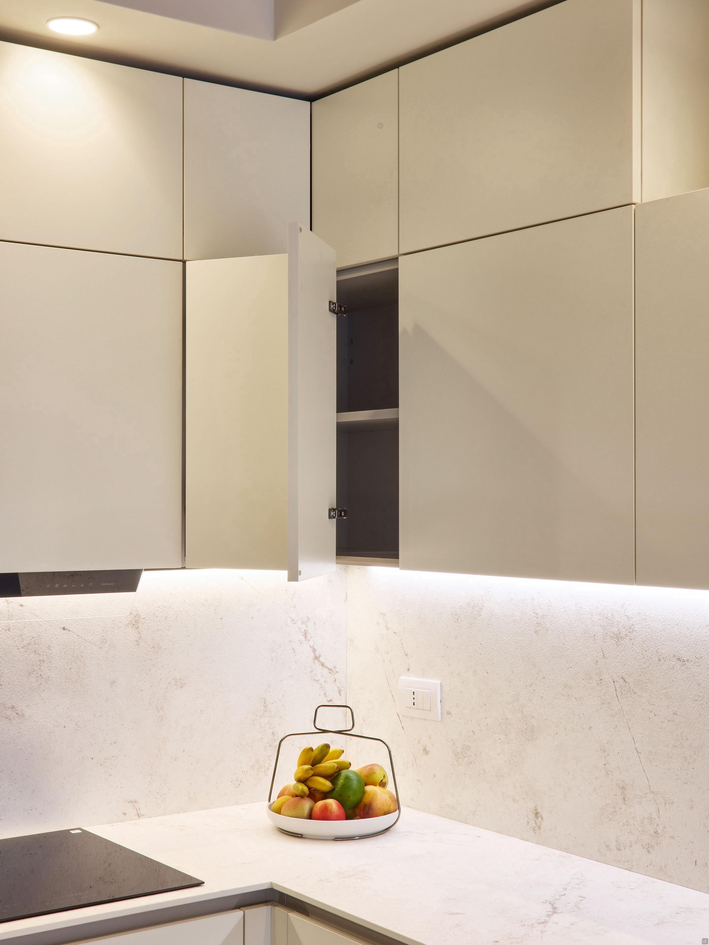 A Modern Kitchens corner by Kitchen Furniture in Yerevan features matte beige custom units. One upper cabinet door is ajar, showing empty shelves, while a white marble countertop holds a wire basket of assorted fruits.